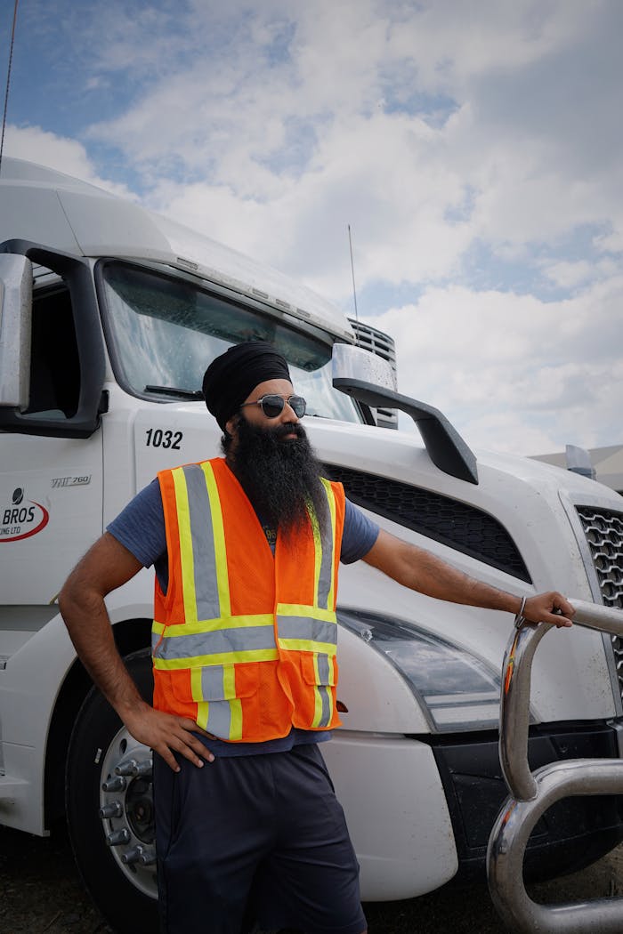 Bearded man in safety vest stands beside a large white truck on a sunny day.