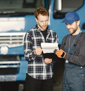 pexels-photo-6720541 Two men in discussion over vehicle maintenance documents in a garage setting with trucks in the background.